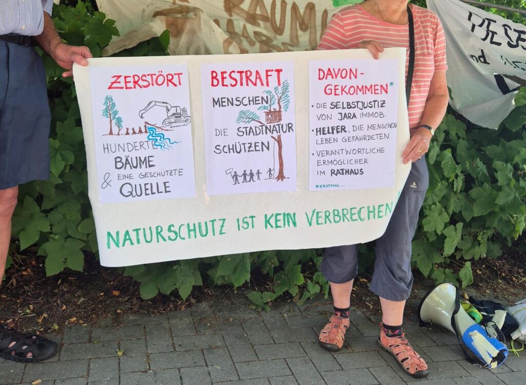 Selbst gemaltes Schild aus drei Teilen "Zerstört - hunderte Bäume & eine geschützte Quelle"
"Bestraft - Menschen, die Stadtnatur schützen"
"Davon gekommen - die Selbstjustiz von Jara Immobilien, Helfer die Menscheleben gefährdeten, verantwortliche Ermöglicher im Rathaus" 
"Naturschutz ist kein Verbrechen" steht groß unten drunter, im Hintergrund ist der Bahnhofswald zu sehen und Menschen, die das Schild halten.