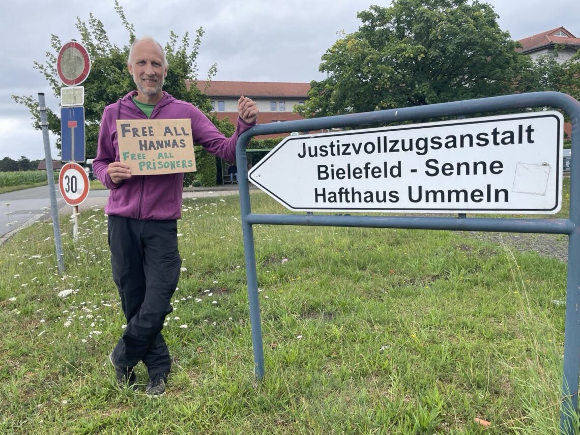 An ein Straßenschild mit der Aufschrift "Justizvollzugsanstalt Bielfeld-Senne, Hafthaus Ummeln" gelehnt steht eine Person, welche ein Schild "Free all Hannas - free all prisoners" hält. Im Hinergrund sind Bäume und ein Gebäude zu sehen.