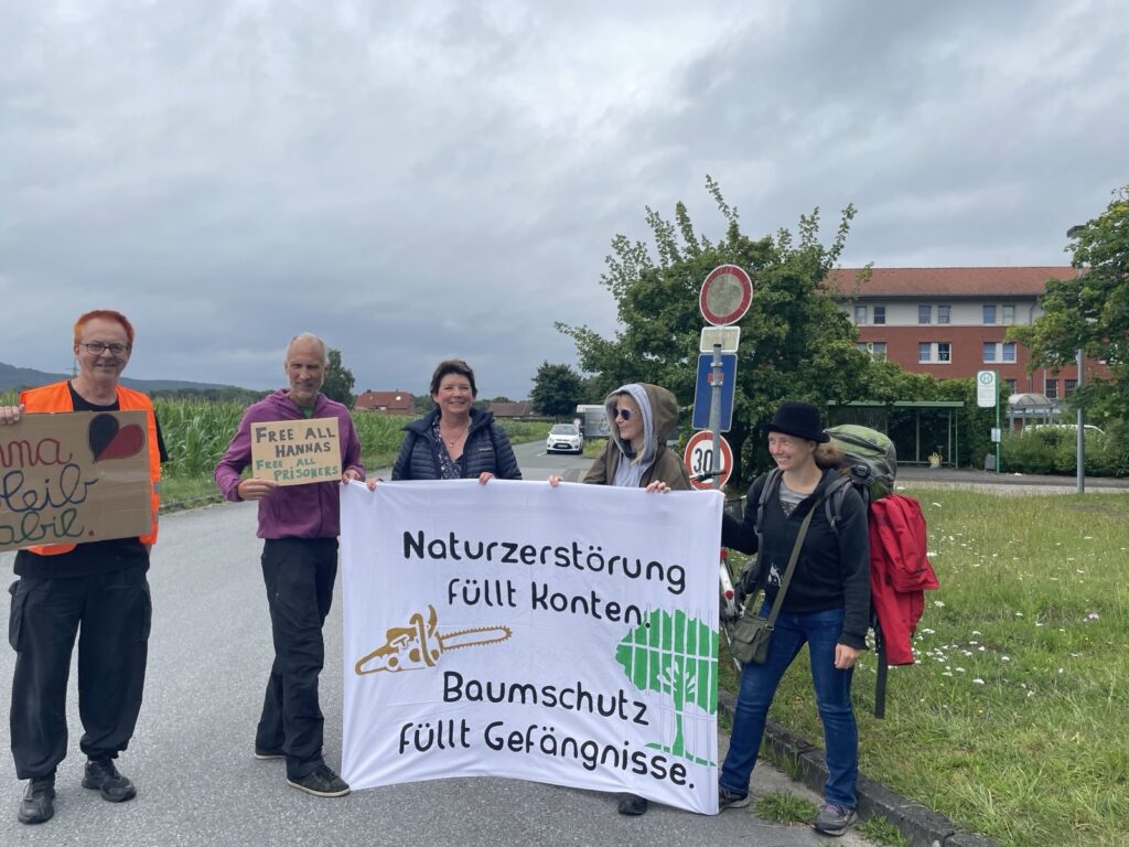 Mehrere Menschen stehen auf einer Straße mit einem Transparent "Naturzerstörung füllt Konten - Baumschutz füllt Gefängnisse". Darauf sind eine Kettensäge und ein Baum hinter Gittern gemalt. Im Hintergrund sind Felder, ein Sackgassenschild, eine Bushaltestelle und ein Haus zu sehen, welches ein Gefängnis sein könnte.