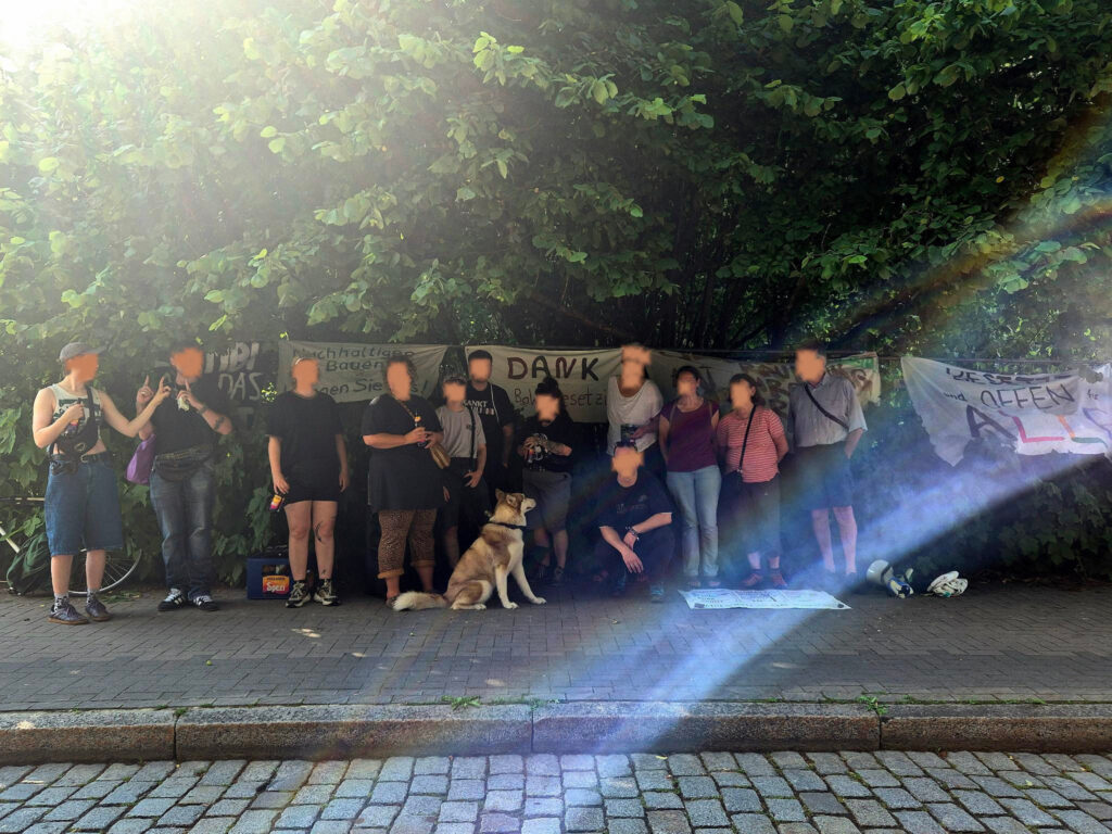 Etliche Menschen und ein Hund stehen vor dem Bahnhofswald aus Solidarität, im Hintergrund sind Transparente zu sehen.